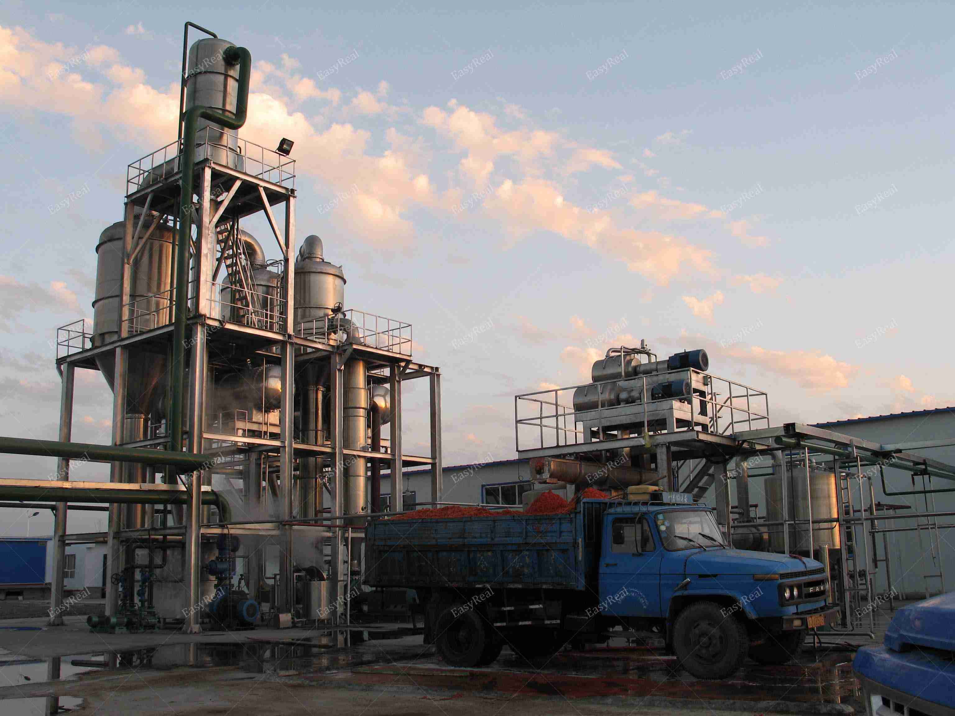Side view of EasyReal forced circulation evaporator structure with platforms and piping, photographed together with a loading truck at the tomato paste processing site.