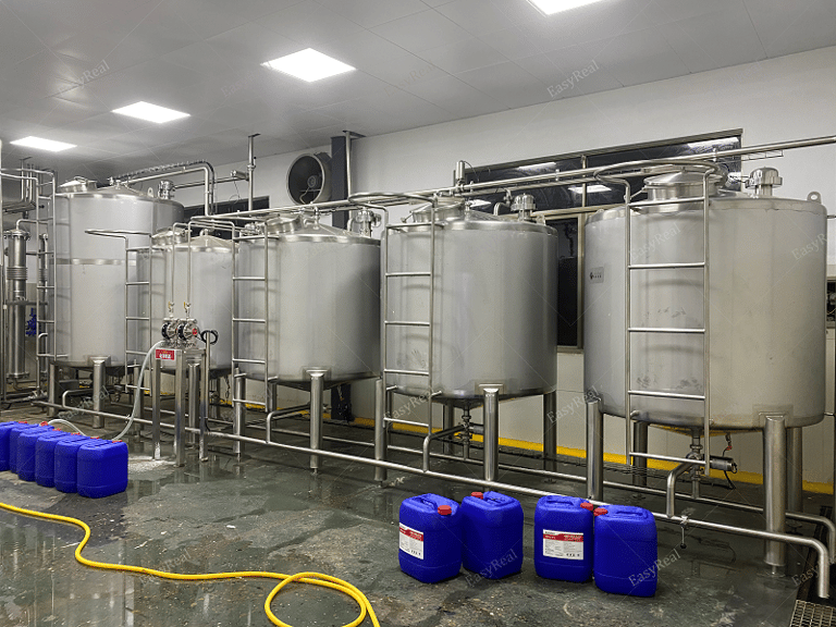 Row of stainless steel mixing and storage tanks with connected process piping and plastic chemical drums on the floor, used for blending, buffering and temporary storage of carrot juice or carrot puree concentrate in the processing plant.