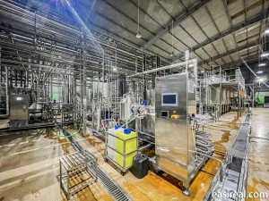Side view of the EasyReal tubular UHT sterilizer integrated into the production line at FGC Vietnam, with the PLC touch-screen control cabinet, tubular heat exchanger sections, CIP chemical containers, and adjacent process equipment and platforms in the beverage workshop.